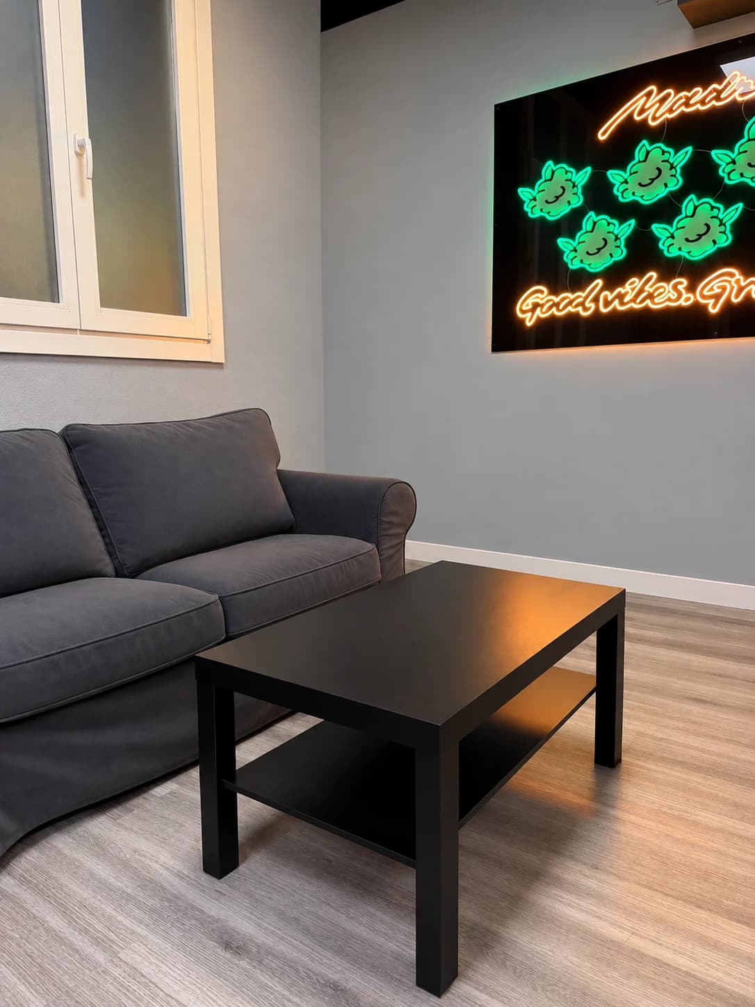 Grey sofa and black table beside the green and orange neon sign at Weed Club Malasaña in Madrid